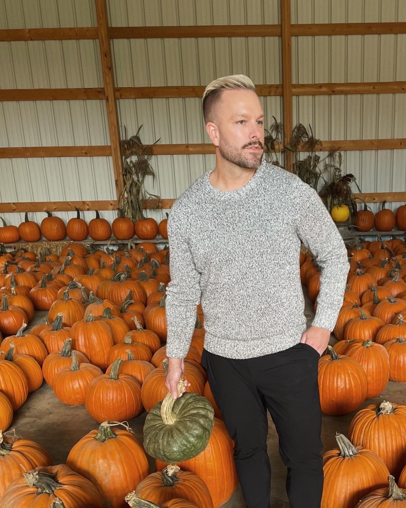 man stands in a pumpkin patch wearing a bulky sweater and black pants