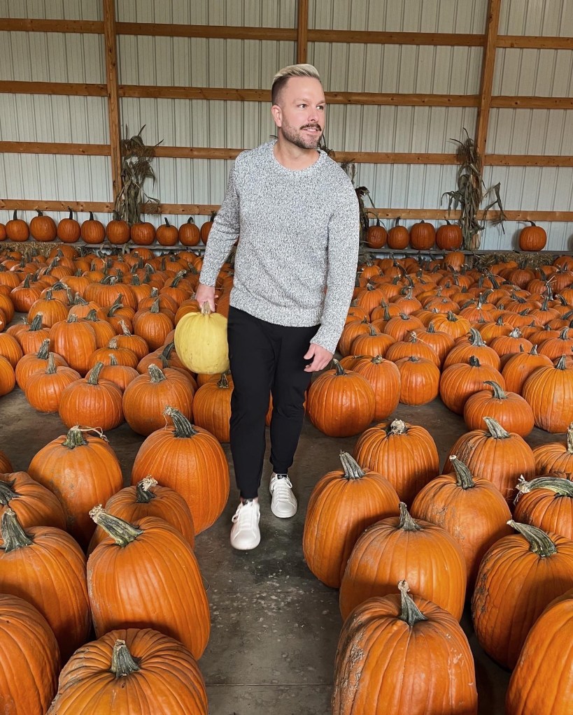 man stands in a pumpkin patch wearing a bulky sweater and black pants