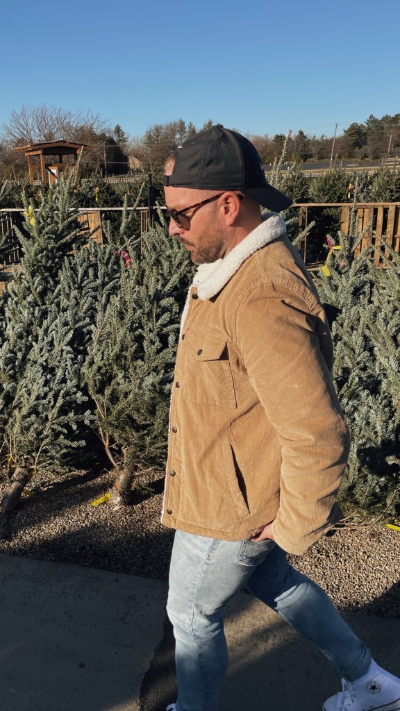 Man walking in front of a row of fresh cut christmas trees. He's wearing a tan levi's trucker jacket and backwards black hat