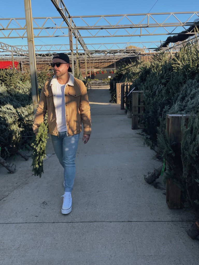 Man walking down a row of christmas trees and wreaths wearing a tan levi's trucker jacket, a backwards black hat, light washed denim and converse high top sneakers